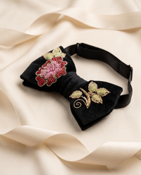 A black velvet bow tie with intricate floral embroidery, featuring a red and pink flower with gold accents on one side and a gold leaf motif on the other, displayed on a draped cream satin fabric.