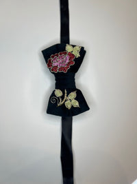 A black velvet bow tie with gold embroidered leaves and a red and pink floral motif on the upper loop, hanging against a plain light background.