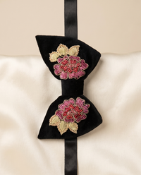 A black velvet bow tie with two embroidered floral motifs in pink, red, and gold, displayed on a cream satin fabric background.