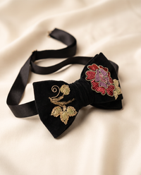 A black velvet bow tie with gold floral embroidery and a red and purple flower accent, displayed on a soft beige fabric background.