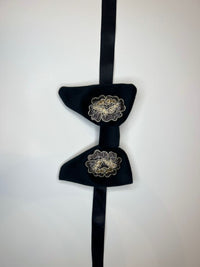 A black velvet bow tie with two gold and white embroidered floral details, hanging against a plain white background.