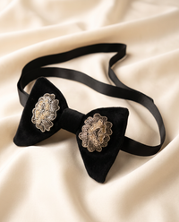 A black velvet bow tie with wide satin neck straps and ornate gold floral appliqué details on each side, displayed on draped cream-colored fabric.