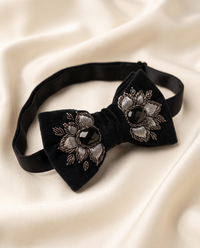 A black velvet bow tie with silver embroidered floral details and dark faceted gemstones, displayed on a draped cream satin fabric background.