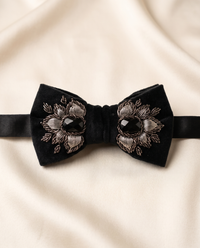 A black velvet bow tie with intricate silver and black floral embroidery and a central black jewel, displayed on a draped cream satin fabric.