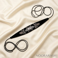 Black velvet Zinat choker necklace with silver floral embroidery and pearl embellishment, laid flat with long cord ties forming loops on draped cream satin fabric.