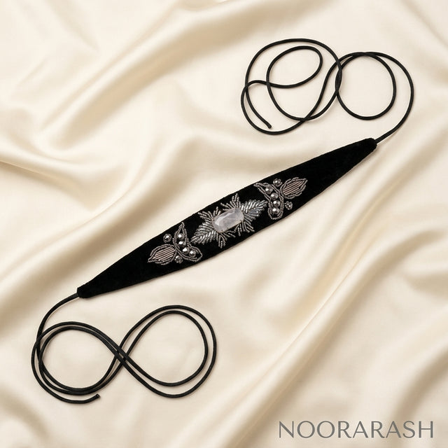 Black velvet Zinat choker necklace with silver floral embroidery and pearl embellishment, laid flat with long cord ties forming loops on draped cream satin fabric.