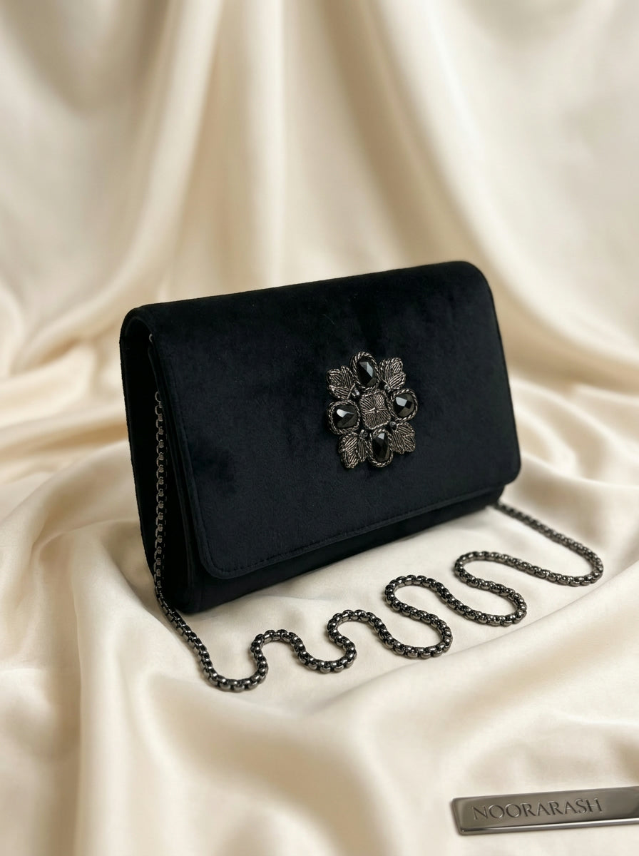 A black velvet clutch bag with a silver-tone floral embellishment and chain strap, displayed on draped cream satin fabric. A brand tag reading NOORASH is visible in the bottom right corner.