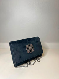 A dark navy blue velvet clutch bag with a silver-tone chain strap and a decorative rhinestone flower clasp, resting on a light-colored surface against a neutral wall.