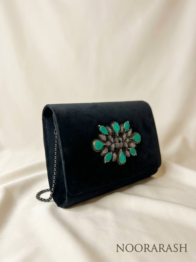 A black velvet clutch bag with a large turquoise and silver floral brooch on the front, resting on a draped cream fabric background. The brand name 'NOORARASH' is printed in the bottom right corner.