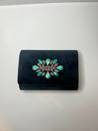 A black velvet clutch purse featuring a decorative brooch on the front flap with turquoise-colored teardrop-shaped stones and a dark metallic center, displayed on a plain light gray surface.