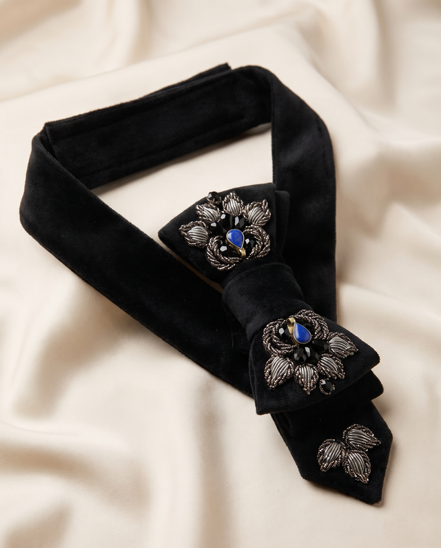 A black velvet bow tie with ornate silver floral embroidery and blue jewel accents, displayed on a draped cream fabric background.