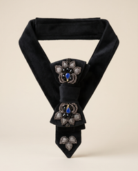 A black velvet necktie with ornate silver-tone floral embroidery and blue jewel accents, displayed on a neutral beige background.