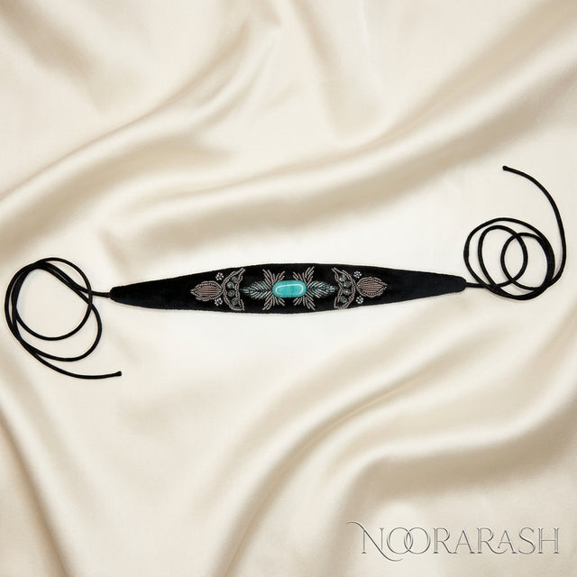 Black velvet Zinat choker necklace with silver and turquoise floral embroidery, laid flat with long cord ties on cream satin fabric.