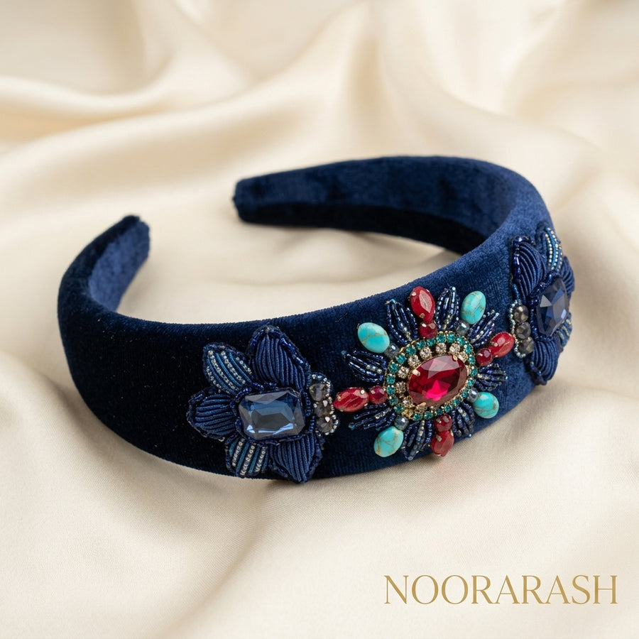 A navy blue velvet headband with intricate floral beadwork and colorful rhinestone embellishments, displayed on a draped cream satin fabric background. The brand name 'NOORARASH' is visible in the bottom right corner.