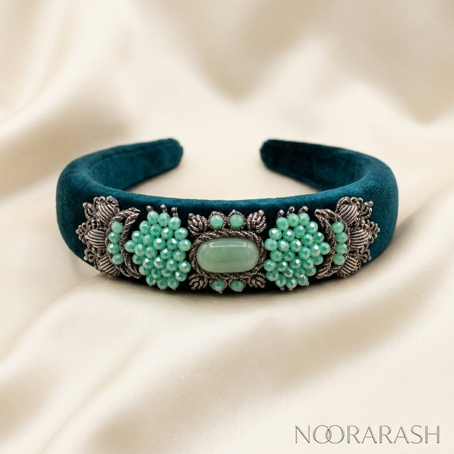 A dark teal velvet cuff bracelet with ornate silver filigree and clusters of mint green beads, displayed on a draped cream satin fabric. The brand name 'NOORARASH' is visible in the bottom right corner.