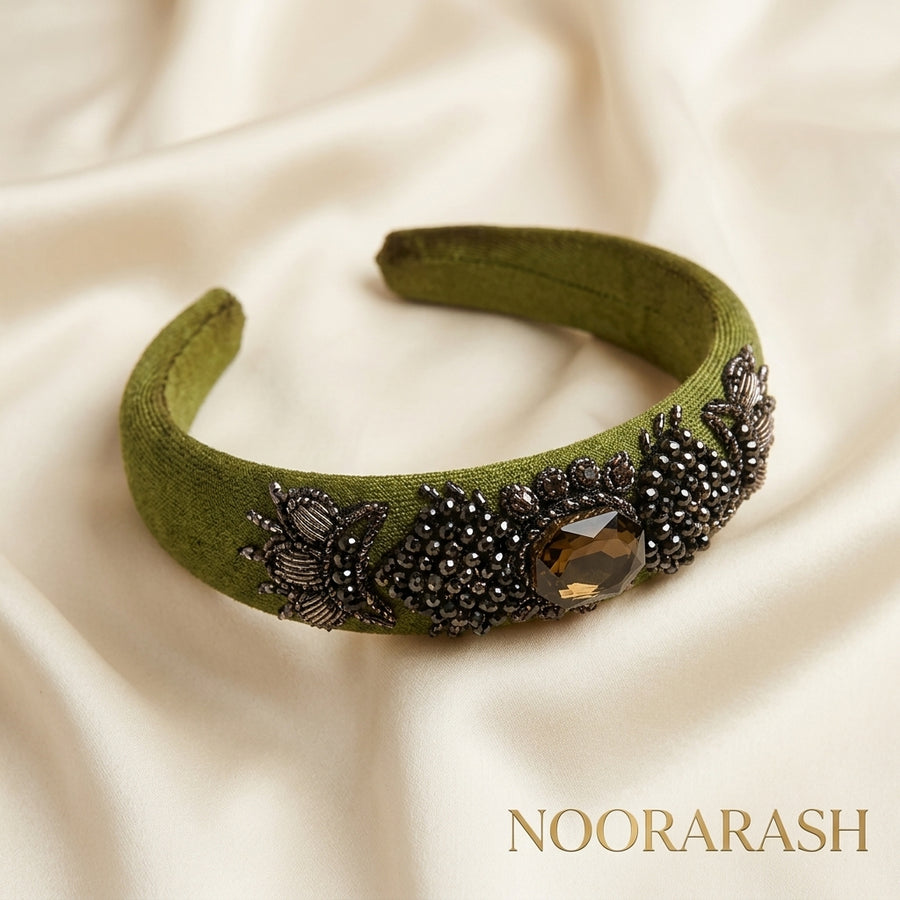 An olive green fabric headband decorated with black beaded floral motifs and a central faceted brown gemstone, displayed on a draped cream satin fabric background. The brand name 'NOORARASH' is printed in gold text in the bottom right corner.