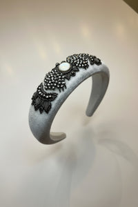 A wide silver fabric headband decorated with black beaded floral patterns and a central white cabochon stone, displayed on a clear glass cup against a light neutral background.