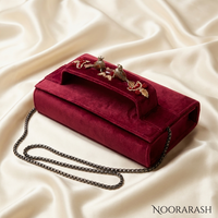 A deep burgundy velvet clutch handbag with a top handle adorned with gold-tone bird embellishments, resting on a draped cream satin fabric background. A dark gunmetal chain strap is draped to the side, and the brand name 'NOORARASH' is visible in the bottom right corner.