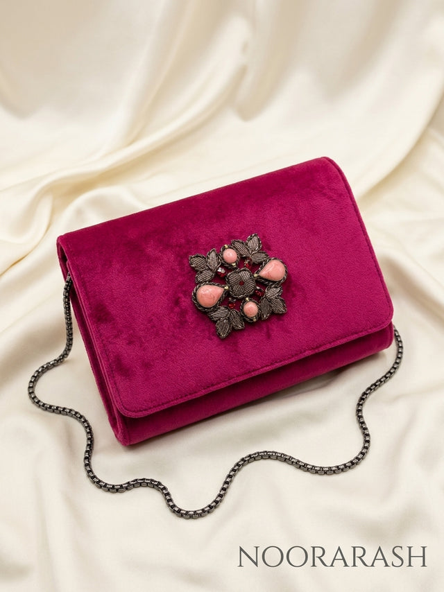 A magenta velvet clutch bag with a dark metal chain strap and a decorative floral embellishment on the front flap, displayed on draped cream satin fabric. The brand name 'NOORARASH' is printed in the bottom right corner.
