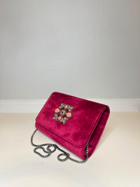 A magenta velvet clutch bag with a decorative floral brooch on the front flap and a silver chain strap, resting on a white surface against a light gray wall.