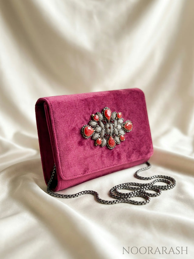 A magenta velvet clutch bag with a silver chain strap and a decorative floral brooch embellished with coral-red stones, displayed on draped white satin fabric.