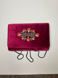 A plum velvet clutch bag with a decorative floral embellishment featuring coral-pink teardrop gems and silver-toned crystals, complete with a black chain strap, shown on a plain off-white background.