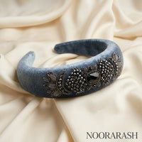 A wide blue velvet headband with ornate silver floral embroidery and a central black rhinestone, displayed on draped cream satin fabric. The brand name NOORARASH is printed in the bottom right corner.
