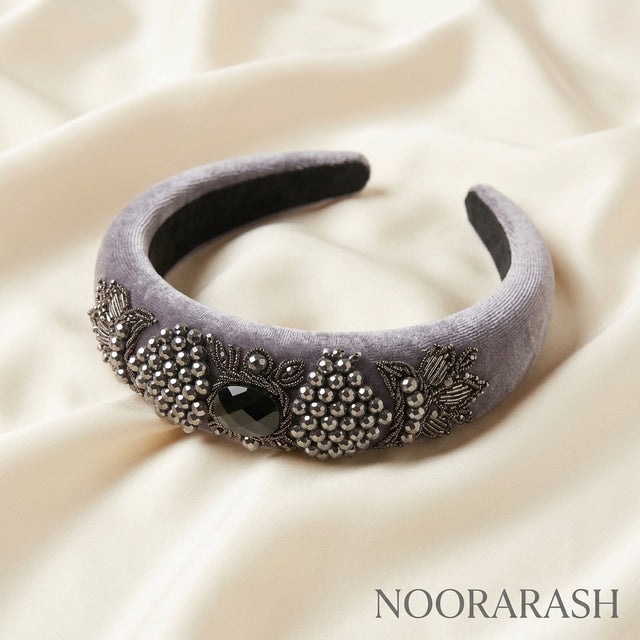 A wide lavender-gray velvet headband decorated with silver bead clusters, floral embroidery, and a central black gemstone, displayed on draped cream satin fabric. The brand name NOORARASH is printed in the bottom right corner.