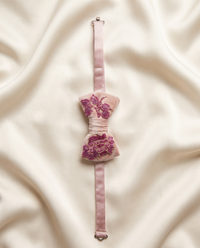 A light pink satin bow tie with purple floral embroidery, laid flat on a draped cream satin fabric background.