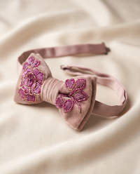 A dusty pink velvet bow tie with intricate magenta and gold floral embroidery on the front, displayed on a soft beige fabric background.