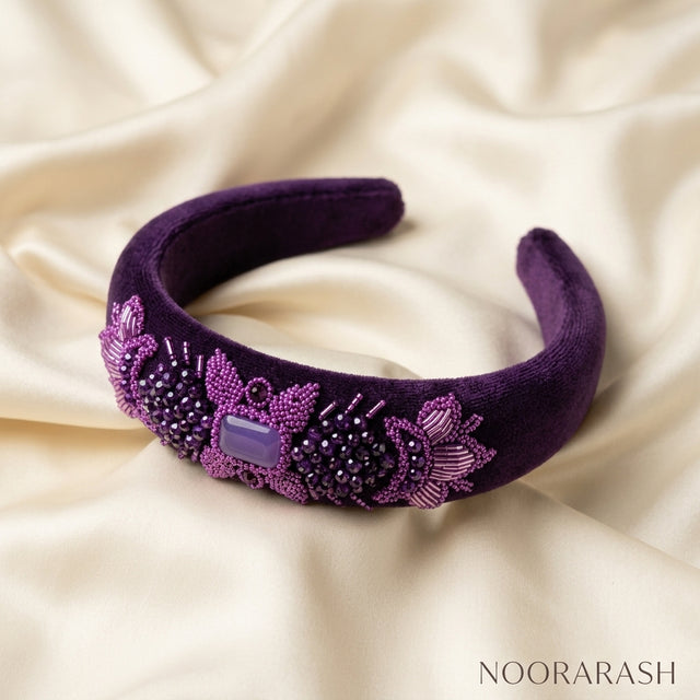A purple velvet headband with intricate floral beadwork and a central purple rectangular stone, displayed on a draped cream satin fabric background. The brand name 'NOORARASH' is visible in the bottom right corner.