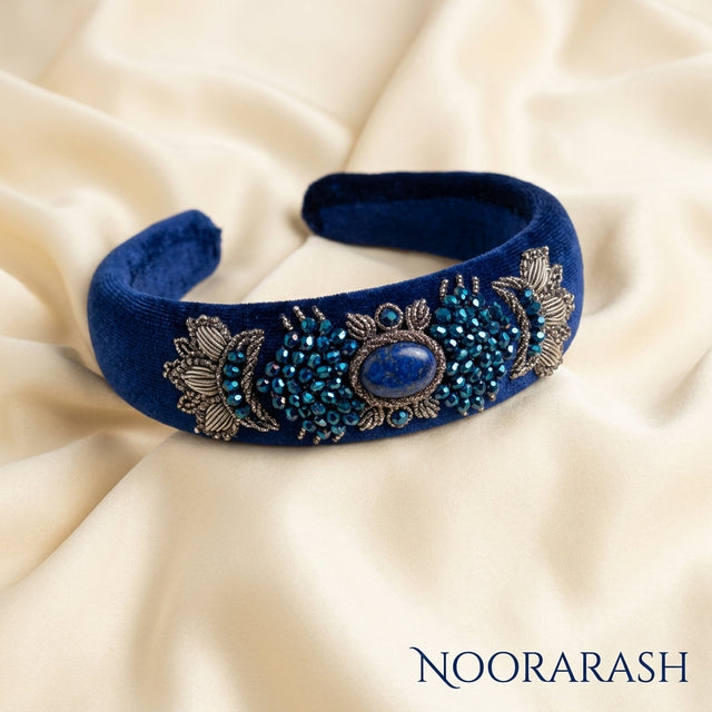 A royal blue velvet headband with intricate silver floral embroidery and blue beadwork, displayed on a draped cream satin fabric background. The brand name 'NOORARASH' is visible in the bottom right corner.