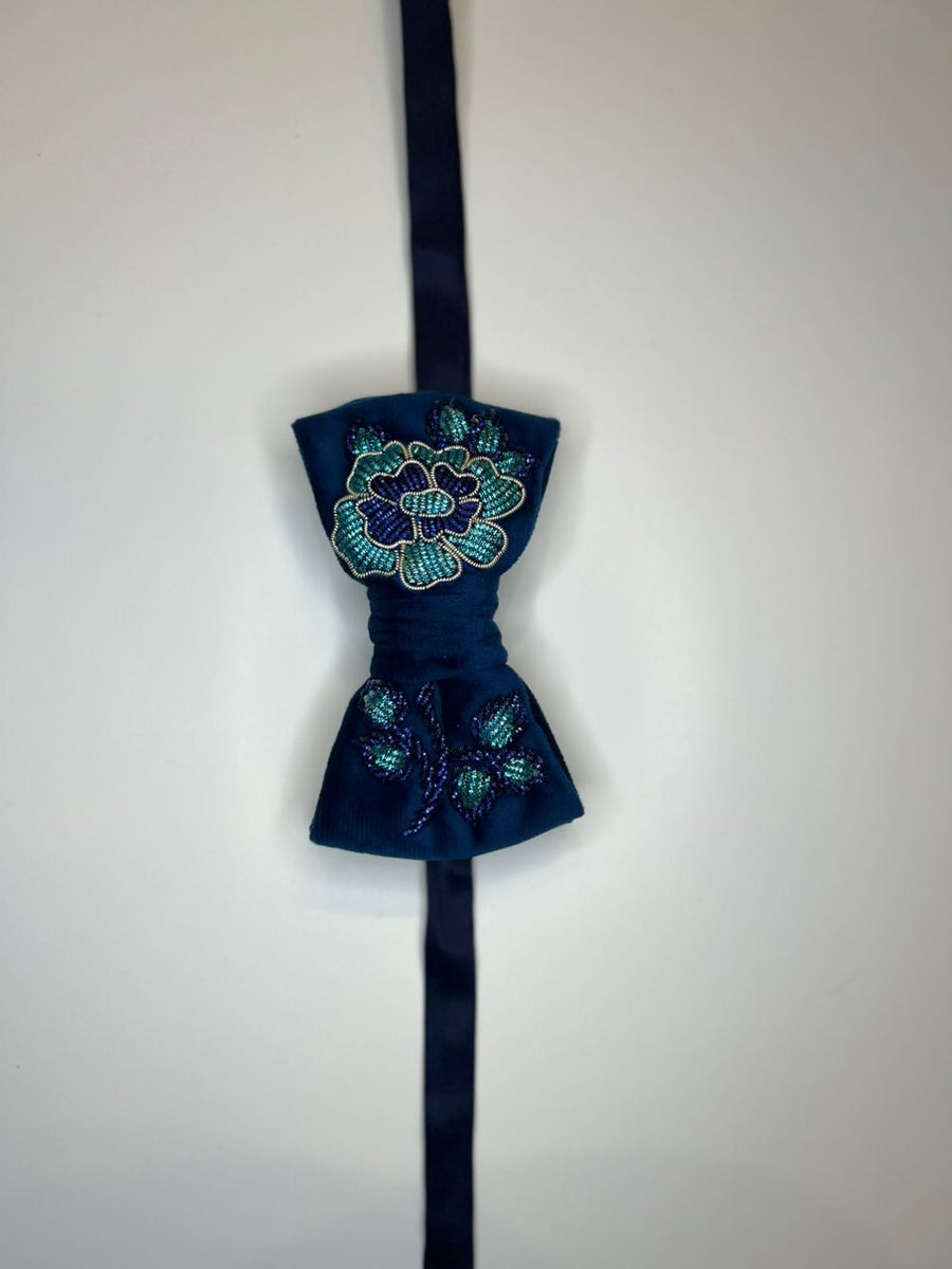 A dark navy blue bow tie with intricate floral embroidery in teal, green, and gold, hanging against a plain off-white background.