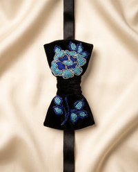 A black velvet bow tie with intricate blue and light blue floral beadwork, displayed on a draped beige satin fabric background.