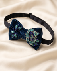 A navy blue velvet bow tie with intricate floral beadwork in green, blue, and gold, displayed on a draped cream satin fabric.
