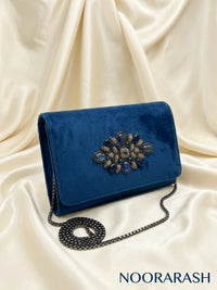 A royal blue velvet clutch bag with a large jeweled floral embellishment on the front flap and a black chain strap, displayed on draped cream satin fabric. The brand name 'NOORARASH' is visible in the bottom right corner.