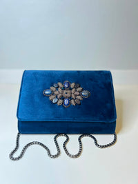 A royal blue velvet clutch bag with a decorative floral brooch featuring blue and silver-toned stones on the front flap, resting on a white surface with a silver chain strap draped in front.