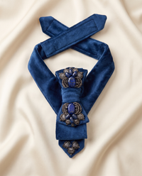 A royal blue velvet necktie with ornate silver-toned filigree brooch details and purple gemstone accents, arranged on a cream-colored satin fabric background.
