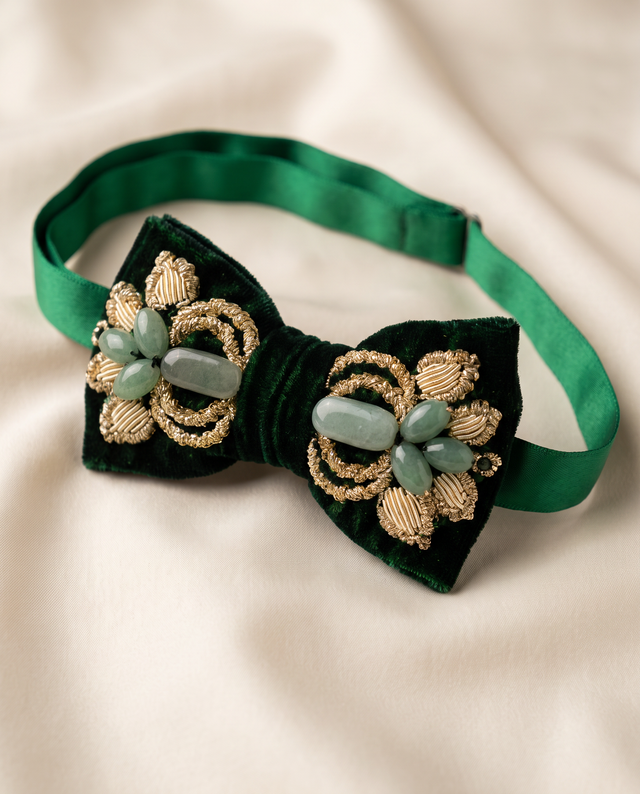 A dark green velvet bow tie with gold embroidered details and light green stone accents, displayed on a soft beige fabric background.