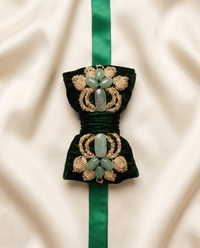 A dark green velvet bow with ornate gold embroidery and light green stone accents, attached to a matching green ribbon on a cream satin background.