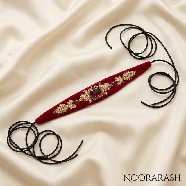 Red velvet Zinat choker necklace with gold floral embroidery and a central brown gemstone, laid flat with long cord ties on cream satin fabric.