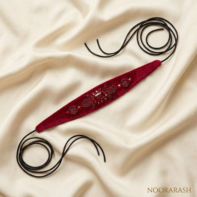 Deep red velvet Zinat choker necklace with intricate beadwork and gold-toned embellishments, laid flat with long cord ties on draped cream satin fabric.