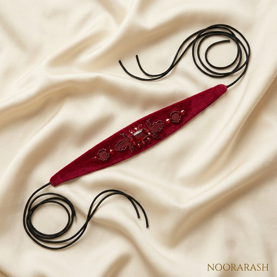 Deep red velvet Zinat choker necklace with intricate beadwork and gold-toned embellishments, laid flat with long cord ties on draped cream satin fabric.